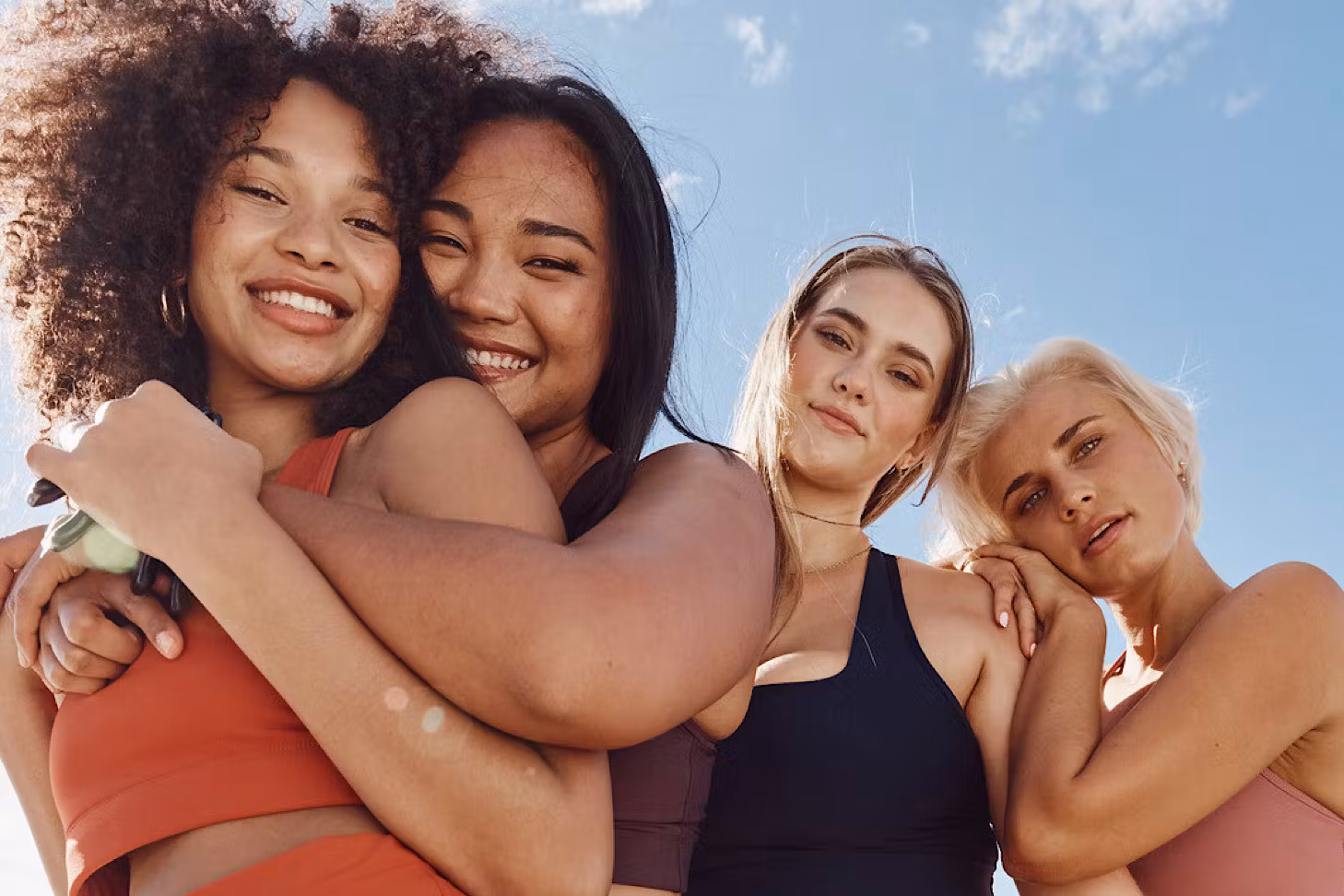 Four friends wear dk.active activewear and hug happily under a blue sky.